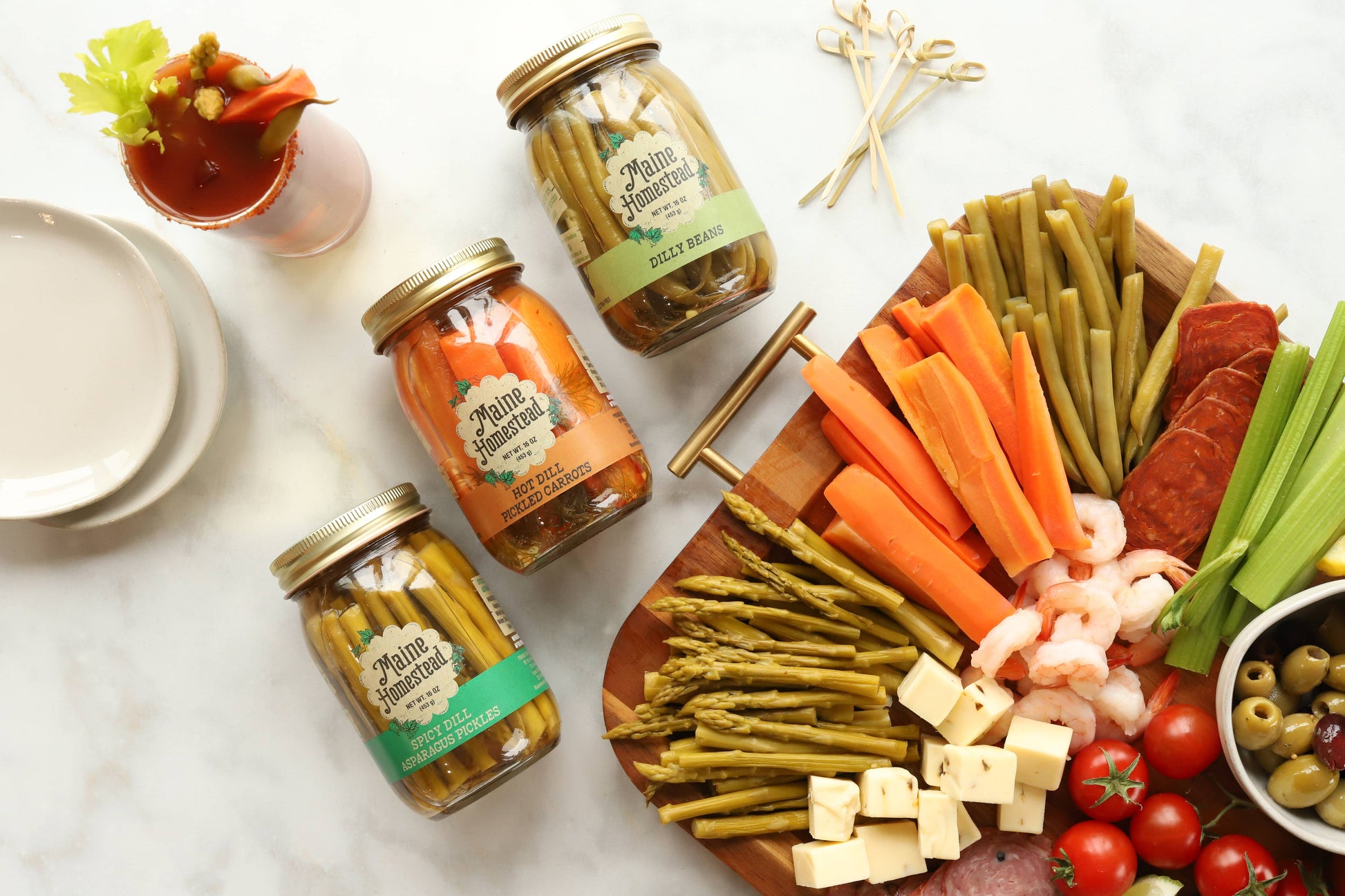Four glass jars of Main Street Pickles arranged on a white marble surface with a styled charcuterie board featuring carrots, asparagus, shrimp, salami, cheese, olives, and tomatoes.