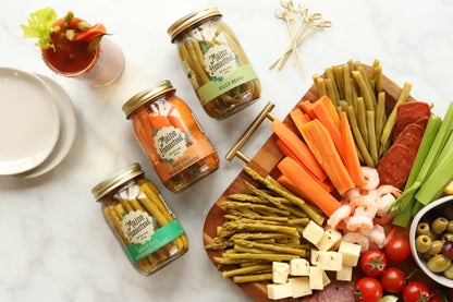 Four glass jars of Main Street Pickles arranged on a white marble surface with a styled charcuterie board featuring carrots, asparagus, shrimp, salami, cheese, olives, and tomatoes.