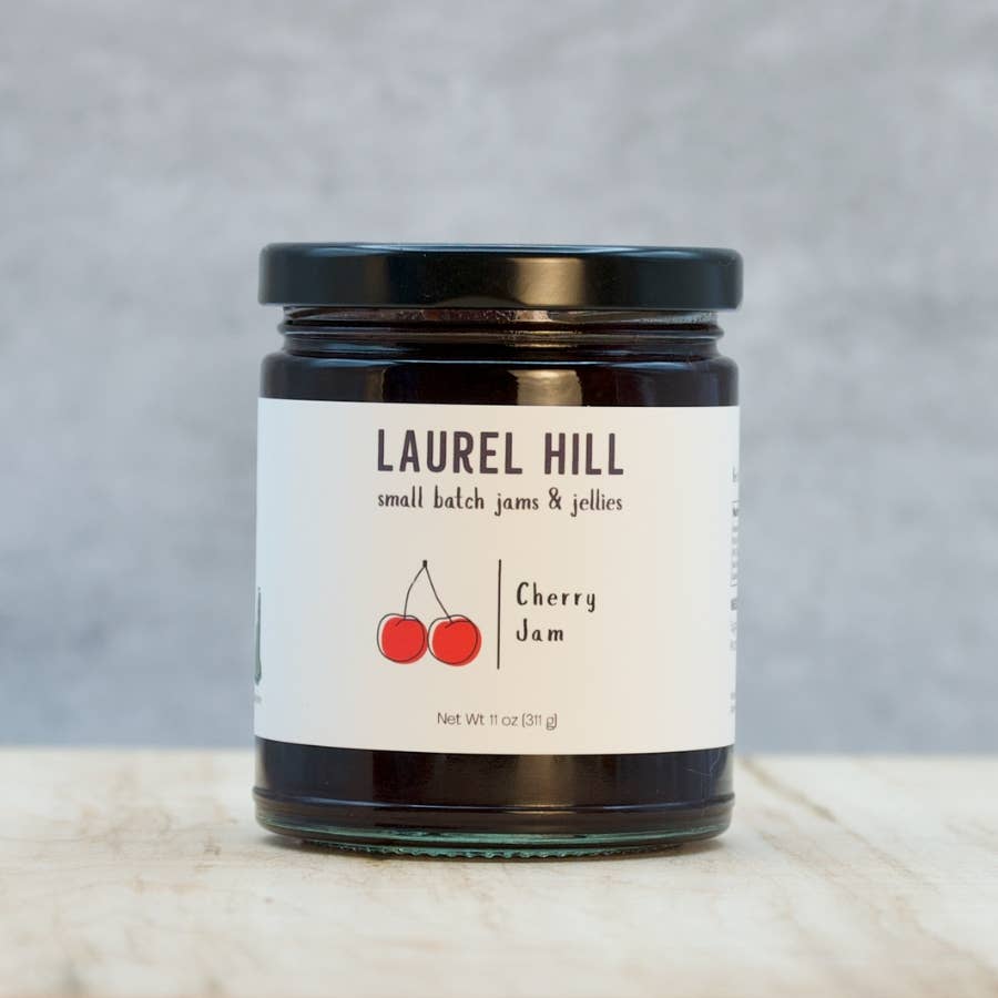 A jar of Laurel Hill Cherry Jam on a light marble surface against a textured gray background. The glass jar has a black lid and a white label with a red cherry illustration.