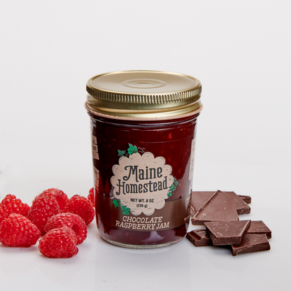 A glass jar of Maine Homestead Chocolate Raspberry Jam with a gold lid, styled with fresh red raspberries on the left and chocolate pieces on the right, on a clean white background.