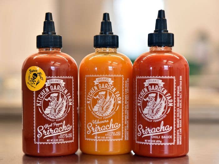 Three bottles of Kitchen Garden Farm Sriracha sauce on a wooden surface with a blurred background.
