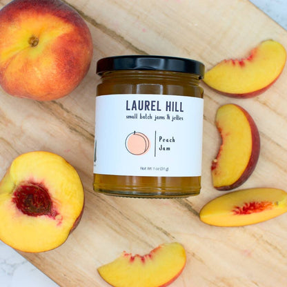 Jar of Laurel Hill Peach Jam surrounded by peaches and slices on a wooden surface