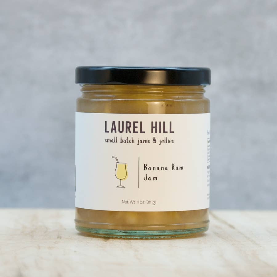 A jar of Laurel Hill Banana Rum Jam sits on a light marble surface against a textured gray background. The glass jar contains golden-brown jam and has a cream-colored label with a black lid.