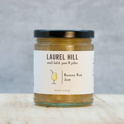 A jar of Laurel Hill Banana Rum Jam sits on a light marble surface against a textured gray background. The glass jar contains golden-brown jam and has a cream-colored label with a black lid.