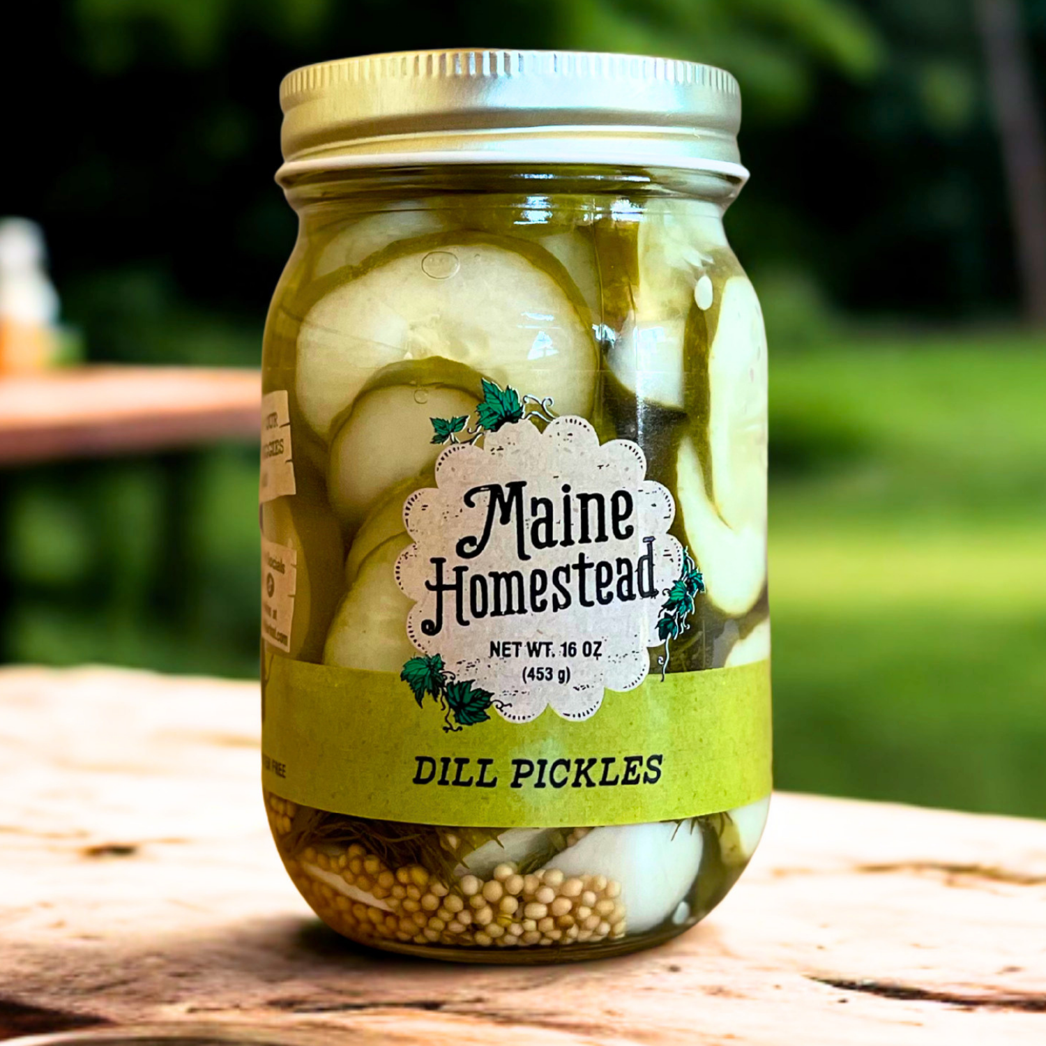 A jar of Maine Homestead dill pickles with sliced dill and onions in olive oil, set on a rustic wooden surface with a blurred green outdoor background.