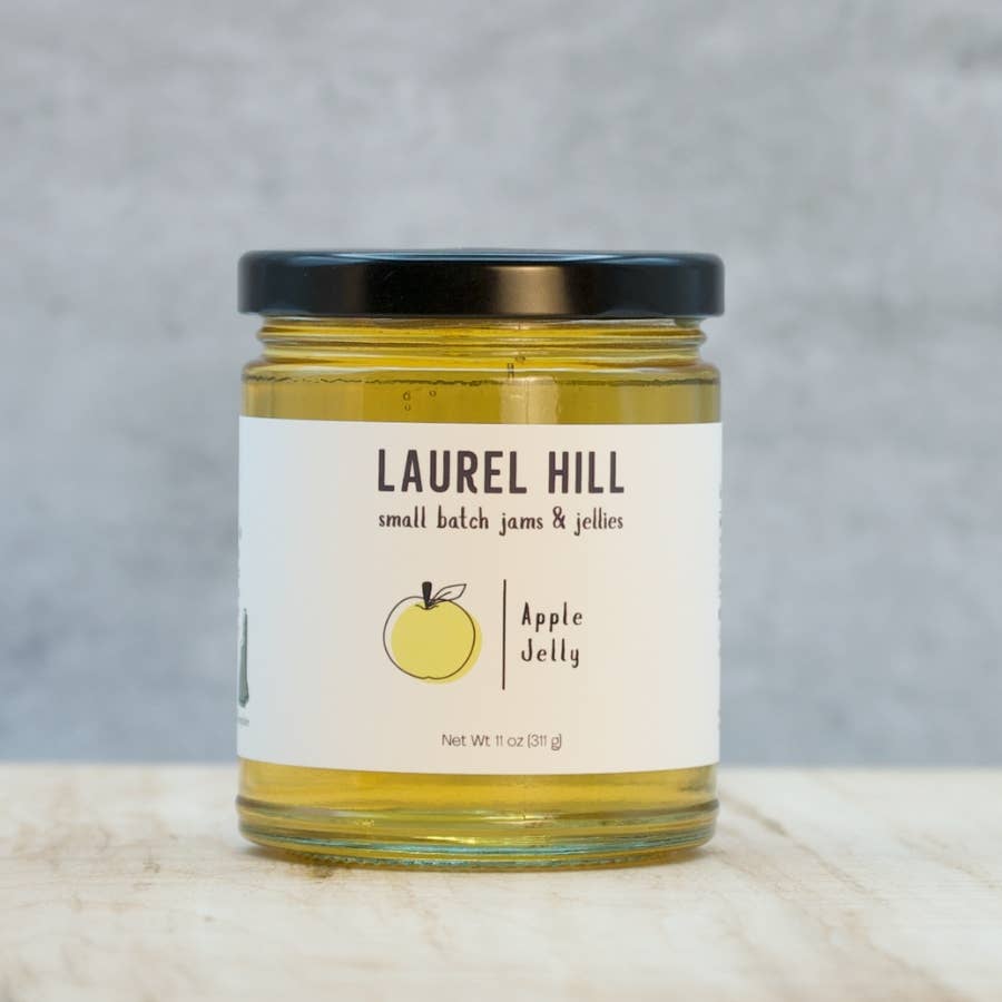 A jar of Laurel Hill Apple Jelly with a black lid and a white label, set on a light wooden surface against a textured gray background.