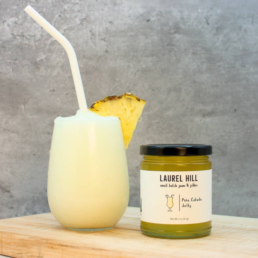 A glass of pale yellow drink with a white straw and pineapple garnish sits beside a small jar labeled Laurel Hill Pine Cactus Juice on a wooden surface against a gray textured wall.