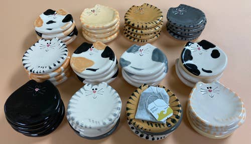 A collection of ceramic cat-shaped coasters and small plates in various colors, including black and white, beige and orange, gray and white, and cream and white, arranged on a solid beige background.
