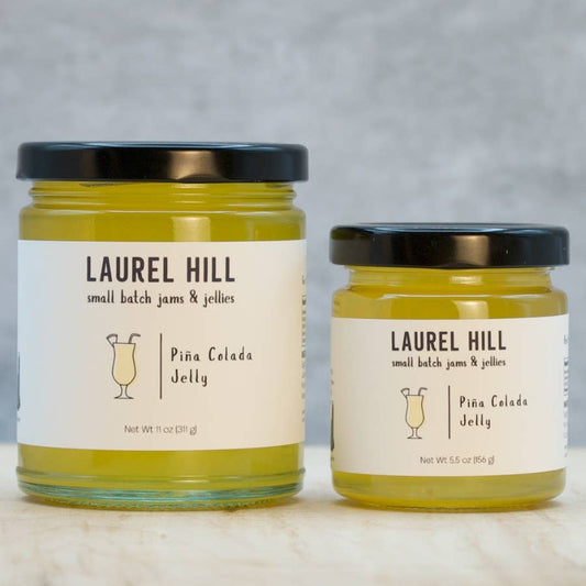 Two glass jars of Laurel Hill Piña Colada Jelly, one 11 oz and one 5.5 oz, with cream labels and black lids, set on a light marble surface against a gray textured background.