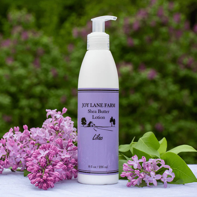 Shea butter lotion bottle with purple label on a white surface with lilac flowers and greenery in the background