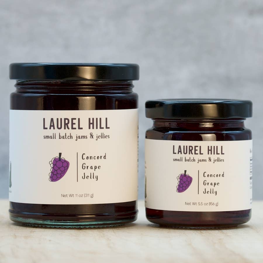 Two glass jars of Laurel Hill Small Batch Jam & Jellies, one larger jar and one smaller jar, both filled with Concord Grape Jelly and featuring white labels with a purple grape illustration, set against a neutral gray background.