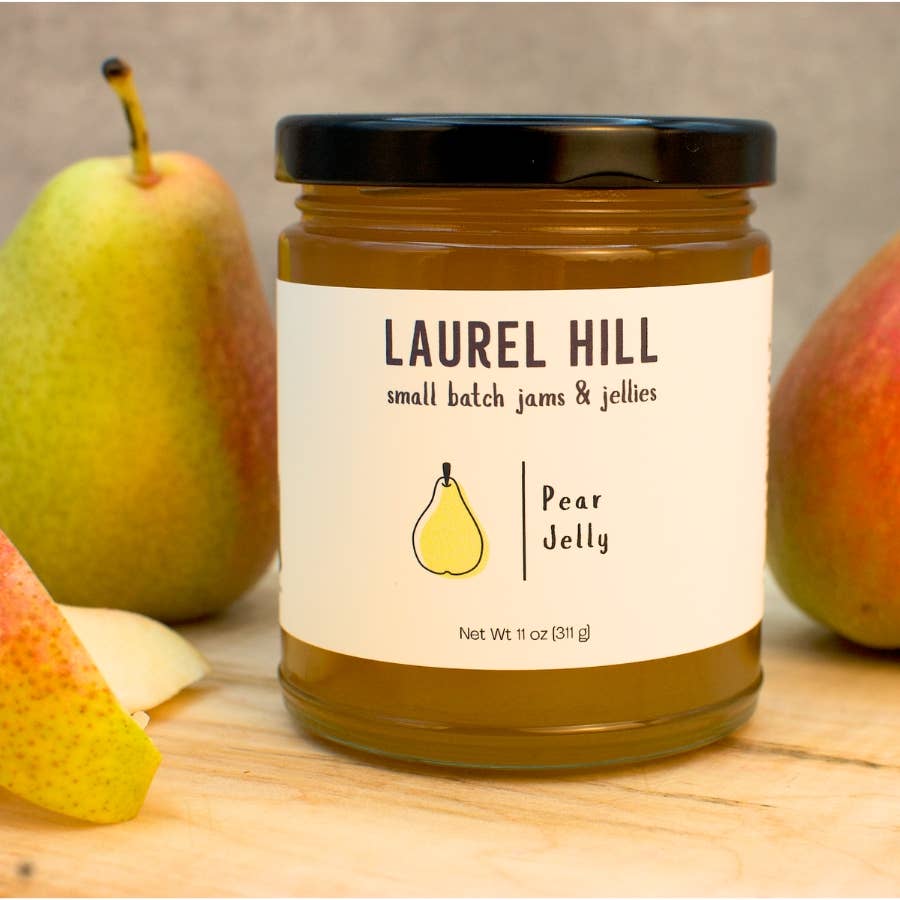 A jar of Laurel Hill Pear Jelly sits on a wooden surface with whole and sliced pears around it. The jar has a black lid and a cream label with the brand name Laurel Hill and a small pear illustration.