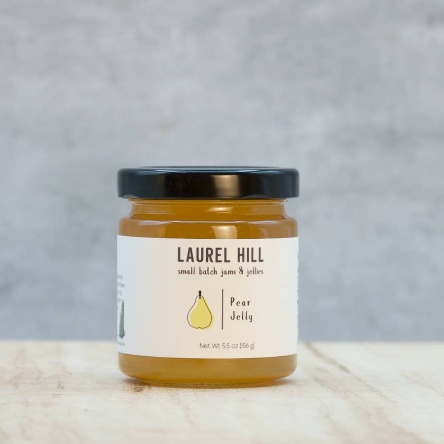 A jar of Laurel Hill Pear Jelly on a light wooden surface with a textured gray background. The glass jar contains golden pear jelly and has a black lid with a white label featuring a pear illustration.