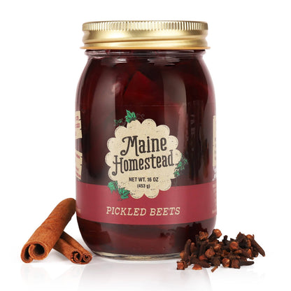 A glass jar of Maine Homestead Pickled Beets with a gold metal lid, set against a white background with whole spices (cinnamon sticks and cloves) placed at its base.