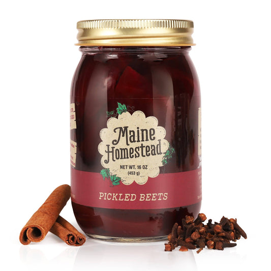 A glass jar of Maine Homestead Pickled Beets with a gold metal lid, set against a white background with whole spices (cinnamon sticks and cloves) placed at its base.