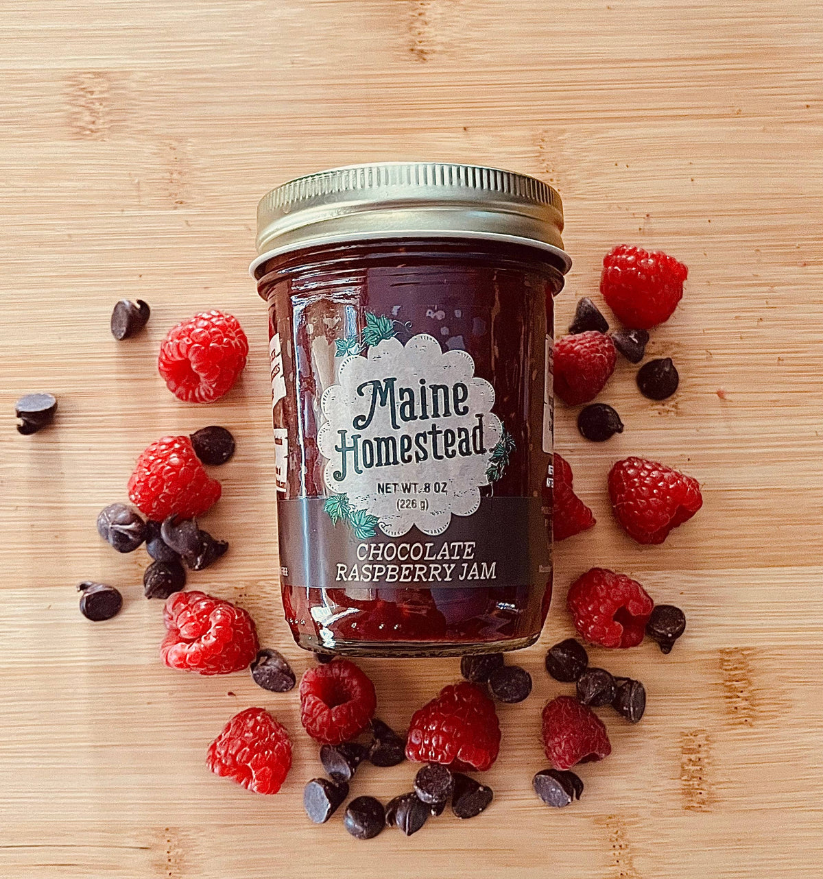 A glass jar of Maine Homestead Chocolate Raspberry Jam sits on a light wood surface, surrounded by fresh raspberries and chocolate chips. The jar has a gold metal lid and a white label with green and black text.