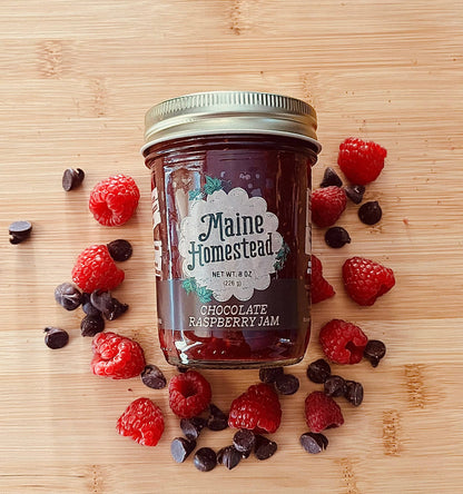 A glass jar of Maine Homestead Chocolate Raspberry Jam sits on a light wood surface, surrounded by fresh raspberries and chocolate chips. The jar has a gold metal lid and a white label with green and black text.