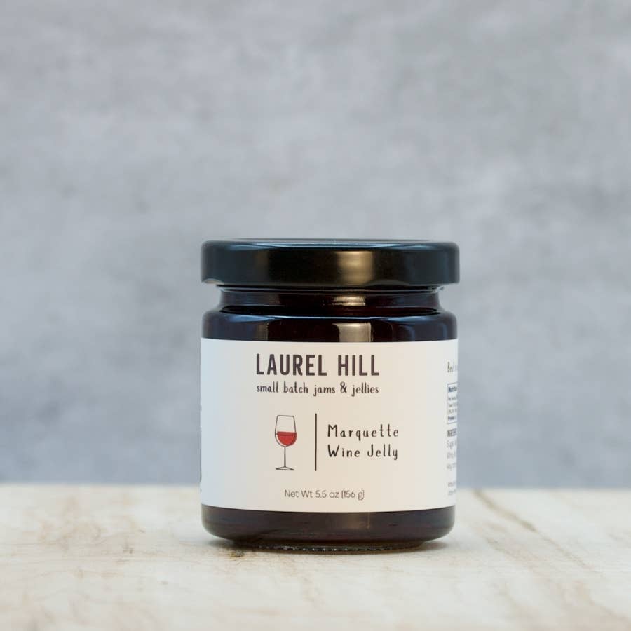 A jar of Laurel Hill Small Batch Jams & Jellies, specifically Marquette Wine Jelly, sitting on a light marble surface against a textured gray background.