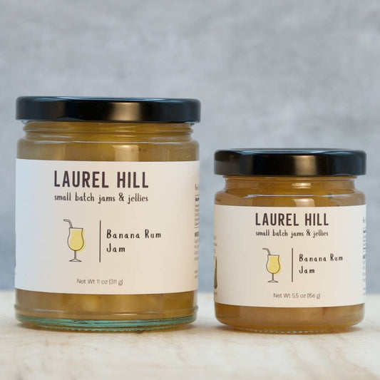 Two glass jars of Laurel Hill Banana Rum Jam with black lids and cream labels, displayed on a light marble surface against a gray background.