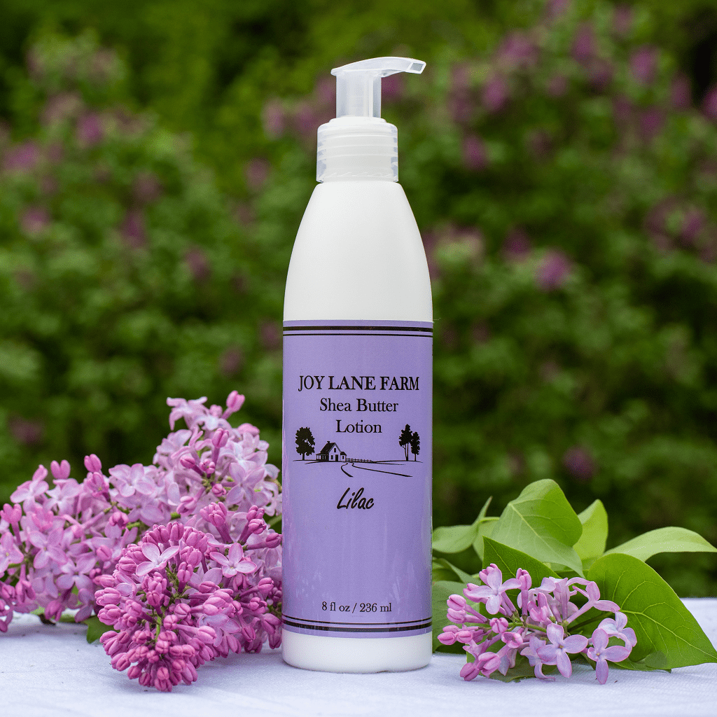 A bottle of Joy Lane Farm Shea Butter Lotion stands on a white surface outdoors, surrounded by pink lilac flowers and green leaves, with a blurred green garden background.