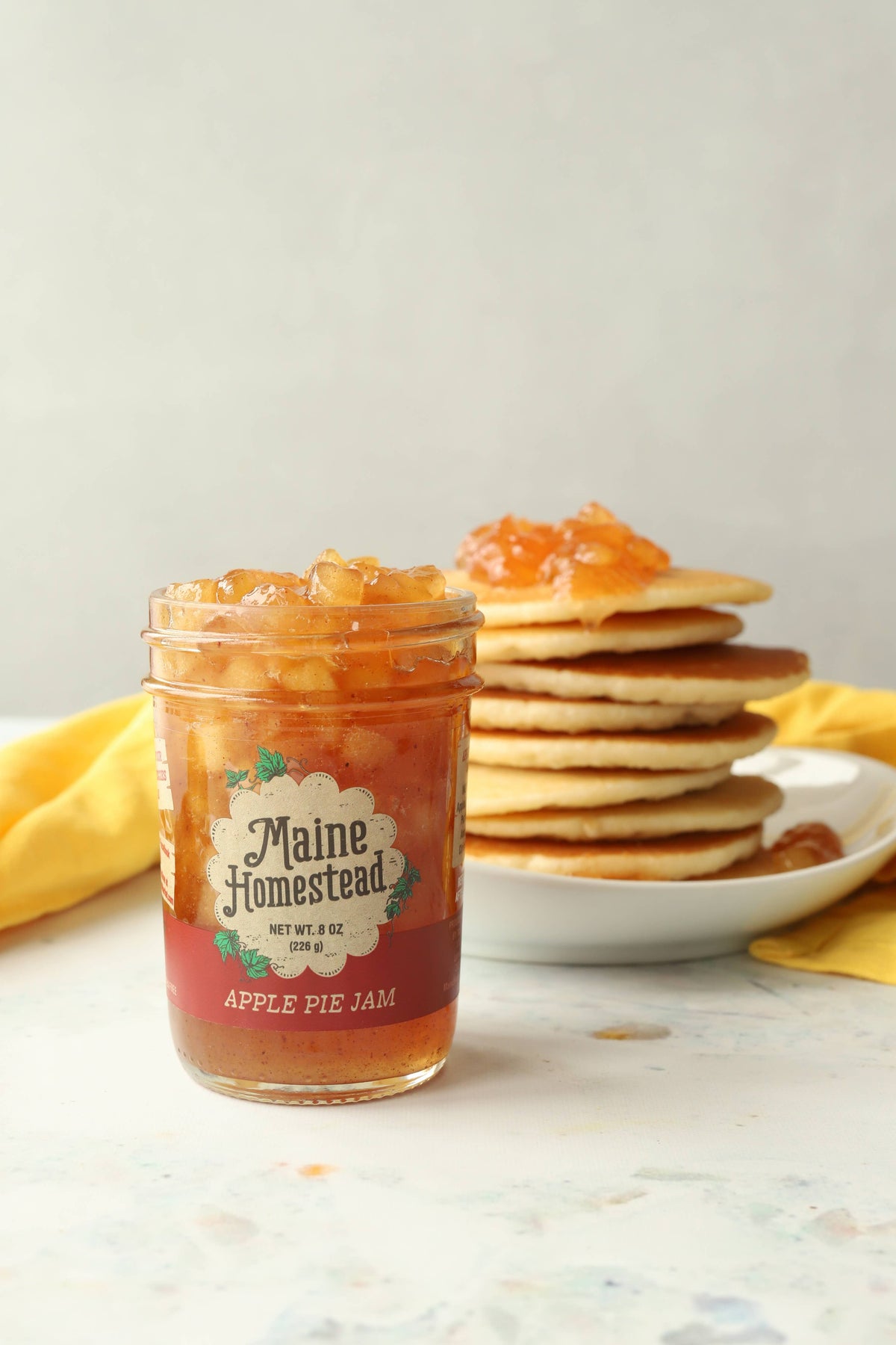 Jar of Maine Homestead Apple Pie Jam with a stack of pancakes on a white surface.