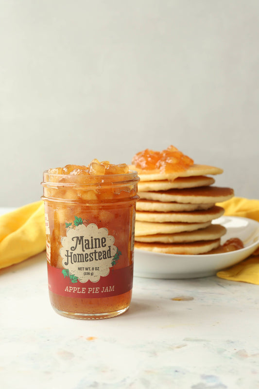 Jar of Maine Homestead Apple Pie Jam with a stack of pancakes on a white surface.