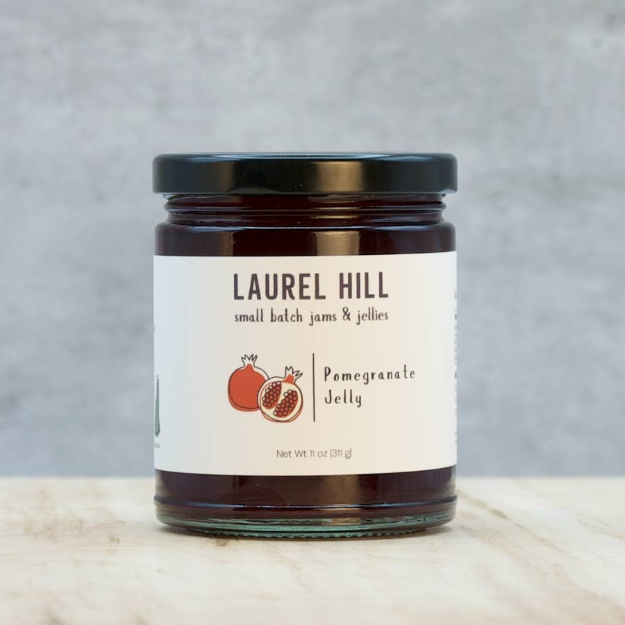 A glass jar of Laurel Hill Small Batch Jams & Jellies pomegranate jelly with a black lid and white label, set on a light marble surface against a textured gray background.