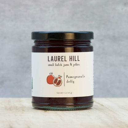 A glass jar of Laurel Hill Small Batch Jams & Jellies pomegranate jelly with a black lid and white label, set on a light marble surface against a textured gray background.