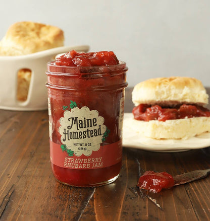 Jar of Maine Homestead strawberry rhubarb jam on a wooden table with biscuits in the background.