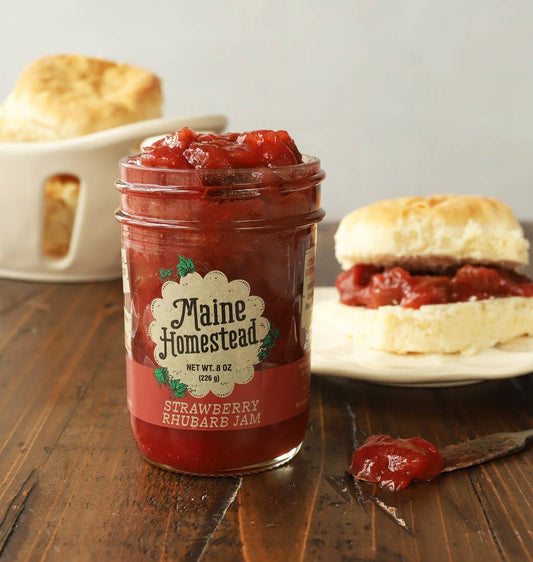 Jar of Maine Homestead strawberry rhubarb jam on a wooden table with biscuits in the background.