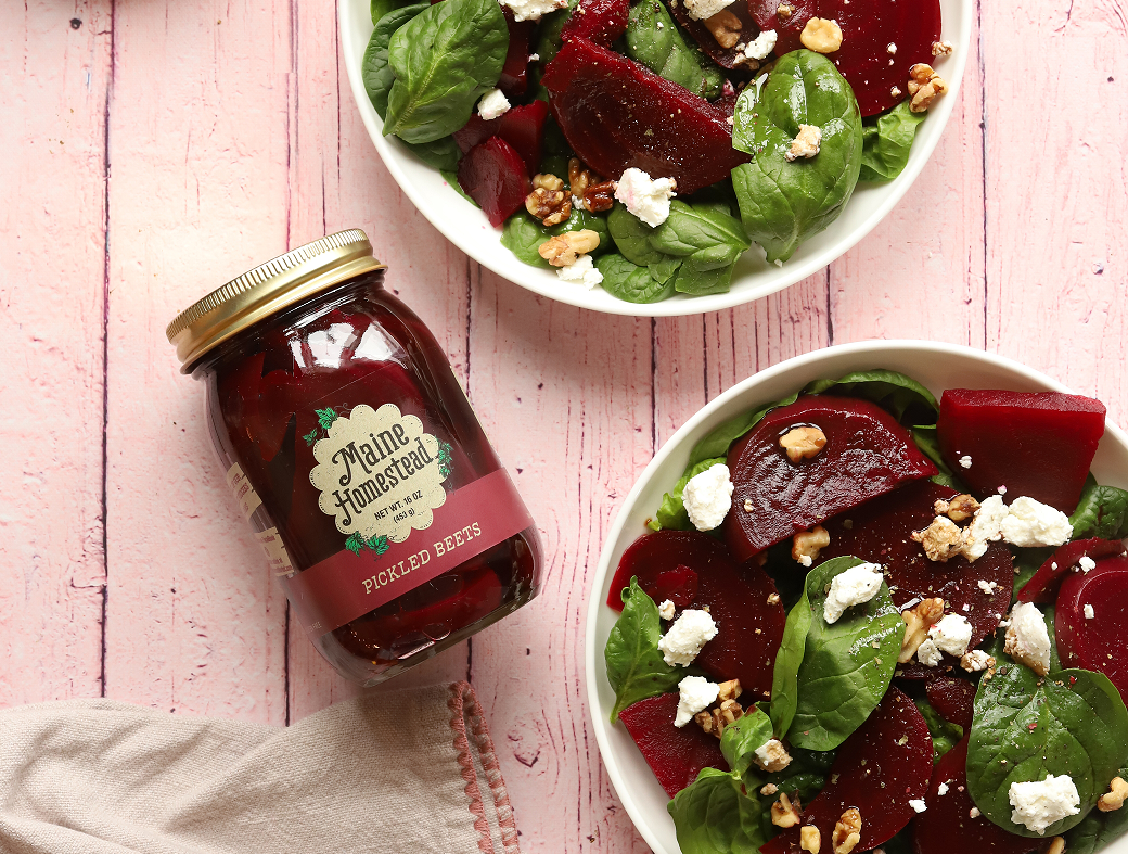 A glass jar of Maine Homesteads Pickled Beets sits on a pink wooden surface next to two white bowls filled with a fresh beet and spinach salad with walnuts and feta cheese.