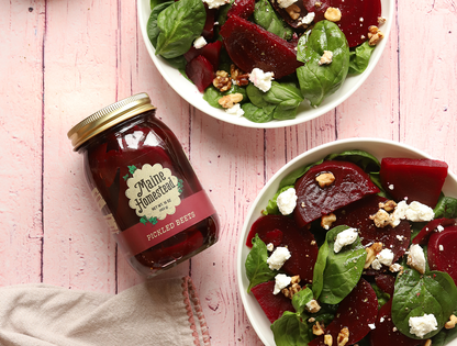 A glass jar of Maine Homesteads Pickled Beets sits on a pink wooden surface next to two white bowls filled with a fresh beet and spinach salad with walnuts and feta cheese.