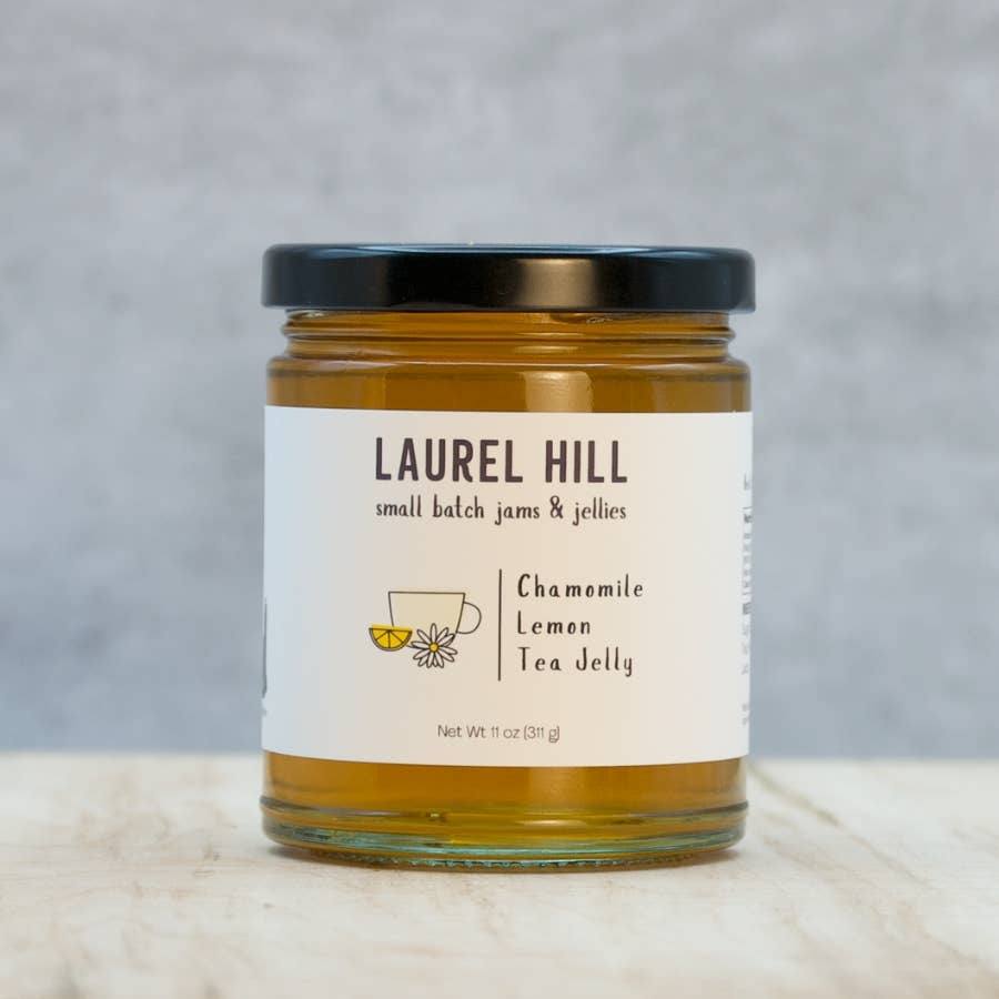 A jar of Laurel Hill Small Batch Jams & Jellies, Chamomile Lemon Tea Jelly, with a white label and black lid, set on a light marble surface against a gray background.