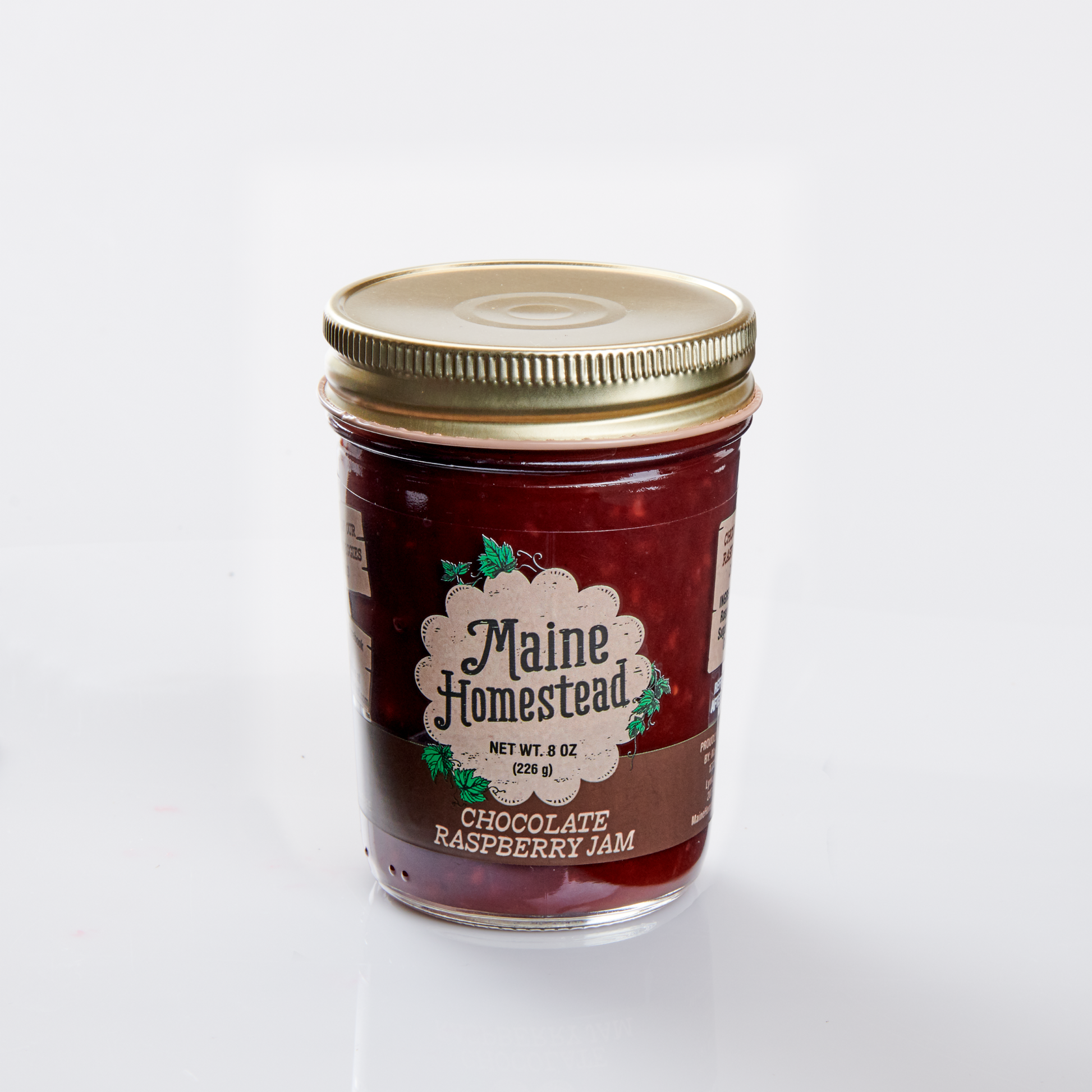 A glass jar of Maine Homestead Chocolate Raspberry Jam with a gold metal lid and a white decorative label, set against a plain white background.