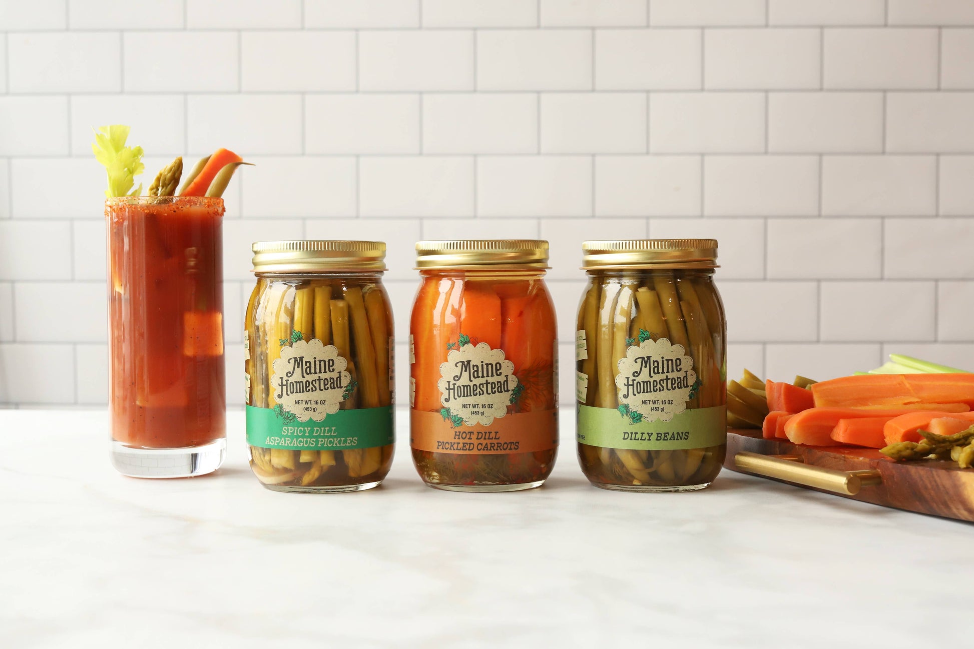 Three glass jars of Maine Seasoned hot and sweet pickle relish are lined up on a white marble countertop with a white subway tile backsplash. A tall glass of red Bloody Mary cocktail with celery and carrot garnish stands to the left, and sliced carrots and celery are visible on a cutting board to the right.