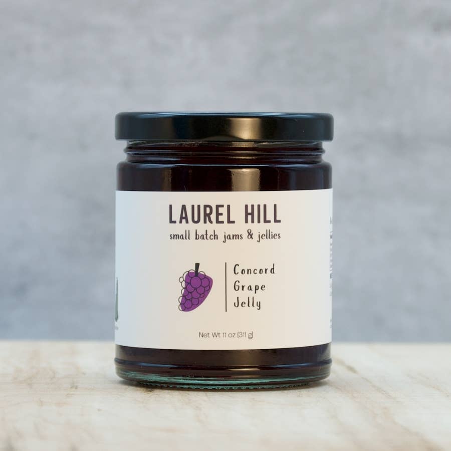 A jar of Laurel Hill Concord Grape Jelly on a light marble surface against a textured gray background. The glass jar contains dark purple jelly, has a black lid, and a white label with a purple grape illustration.