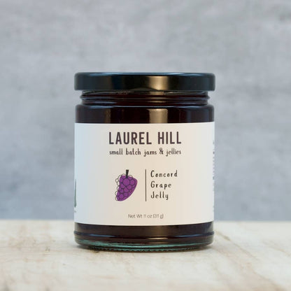 A jar of Laurel Hill Concord Grape Jelly on a light marble surface against a textured gray background. The glass jar contains dark purple jelly, has a black lid, and a white label with a purple grape illustration.