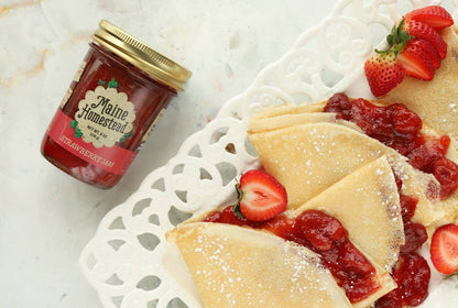 A glass jar of Maine Homestead brand strawberry jam sits next to a white decorative plate filled with folded crepes topped with fresh strawberry slices and jam, on a light marble surface.