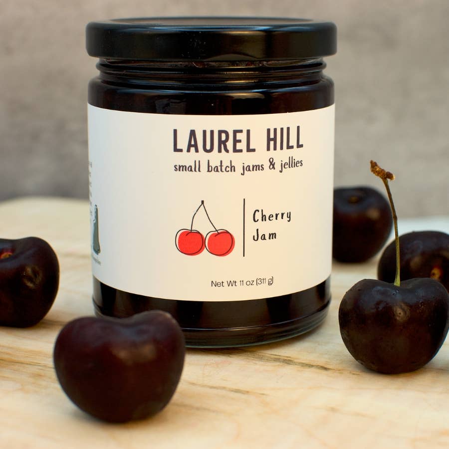 A jar of Laurel Hill Cherry Jam sits on a light wooden surface with fresh dark cherries placed around it. The jar has a black lid and a white label with the brand name Laurel Hill and a red cherry illustration.