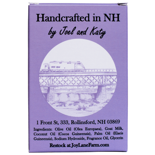 A rectangular purple product box with printed text reading 'Handcrafted in NH by Joel and Katy' and a circular illustration of a train crossing a bridge. The box also includes an address and ingredient list, plus 'Restock at JoyLaneFarm.