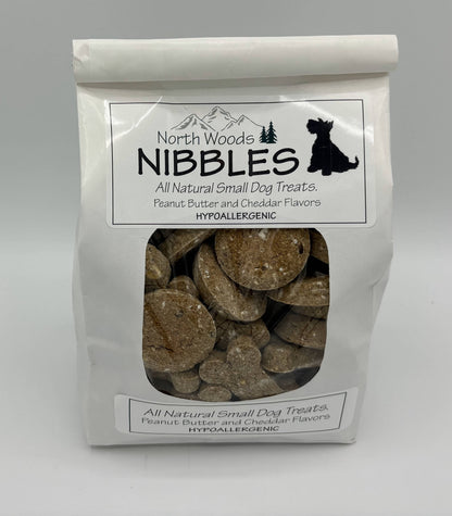 A white stand-up pouch of North Woods Nibbles All Natural Small Dog Treats, featuring a clear window that displays round, tan dog treats inside. The packaging includes a black dog silhouette logo and text indicating peanut butter and cheddar flavors.