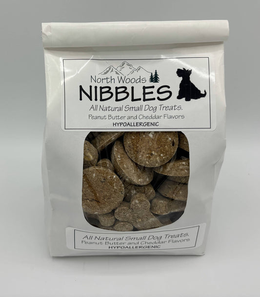 A white stand-up pouch of North Woods Nibbles All Natural Small Dog Treats, featuring a clear window that displays round, tan dog treats inside. The packaging includes a black dog silhouette logo and text indicating peanut butter and cheddar flavors.