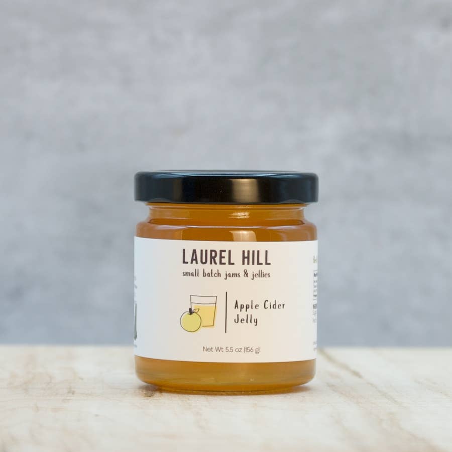 A jar of Laurel Hill Apple Cider Jelly on a light marble surface with a textured gray background. The jar has a black lid and a white label with a small yellow apple illustration.
