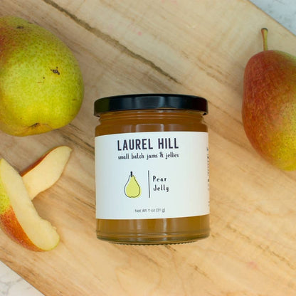 A jar of Laurel Hill Pear Jelly sits on a wooden cutting board surrounded by fresh pears and pear slices. The glass jar has a black lid and a white label with a small yellow pear illustration.
