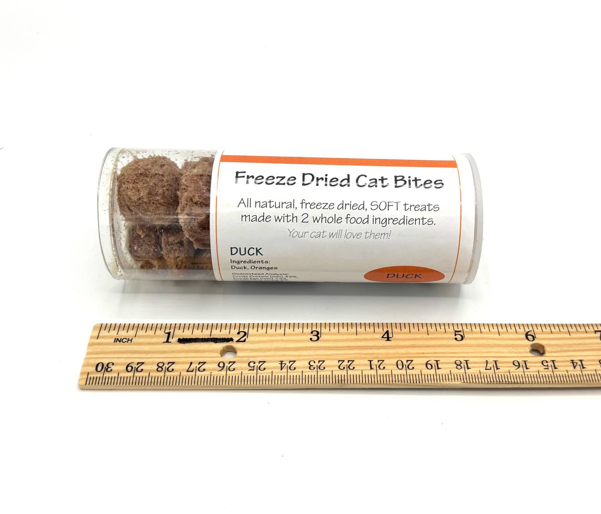 A clear plastic tube of freeze dried cat bites treats with a white label, shown next to a wooden ruler on a white background. The label reads 'Freeze Dried Cat Bites' and 'DUCK'.