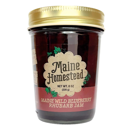 A jar of Maine Homestead Maine Wild Blueberry Rhubarb Jam with a gold metal lid and a cream and red label, set against a plain white background.