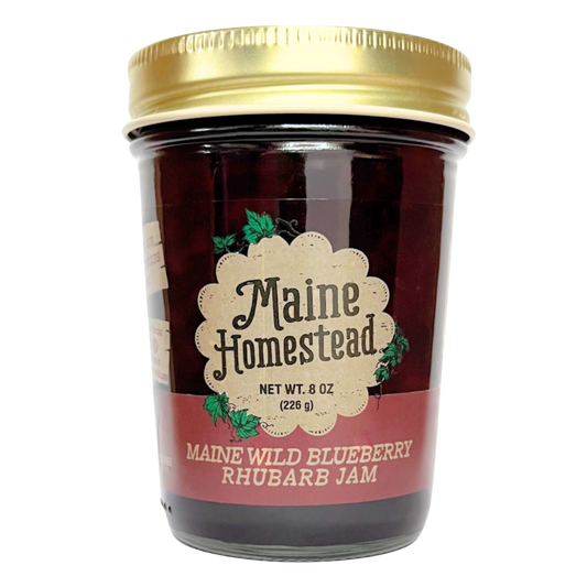 A jar of Maine Homestead Maine Wild Blueberry Rhubarb Jam with a gold metal lid and a cream and red label, set against a plain white background.