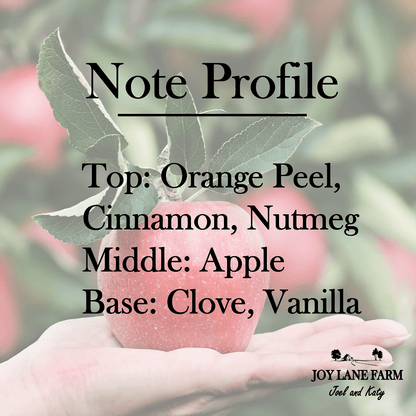 A hand holds a small red apple with green leaves against a blurred apple tree background. Large black text reads 'Note Profile' with scent notes: 'Top: Orange Peel, Cinnamon, Nutmeg', 'Middle: Apple', and 'Base: Clove, Vanilla'.