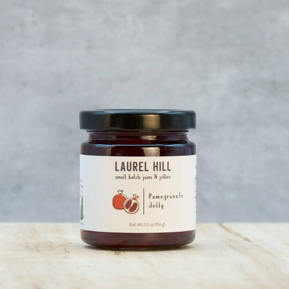 A jar of Laurel Hill Pomegranate Jelly sits on a light marble surface against a textured gray background. The product is in a dark glass jar with a black lid and a cream-colored label featuring red pomegranate illustrations.