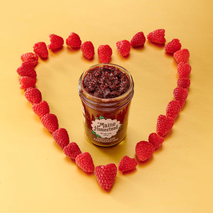 A jar of Maine Homestead raspberry jam is centered on a yellow background, surrounded by a heart shape made of fresh raspberries. The jar label reads 'Maine Homestead'.